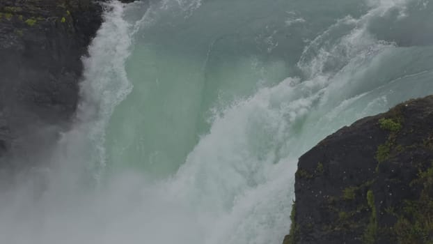 Slow-mo video shows a glacial waterfall's vibrant turquoise flow amid a soft rainfall.
