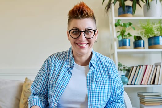 Portrait of happy smiling middle-aged woman in glasses with red haircut looking at camera in home interior