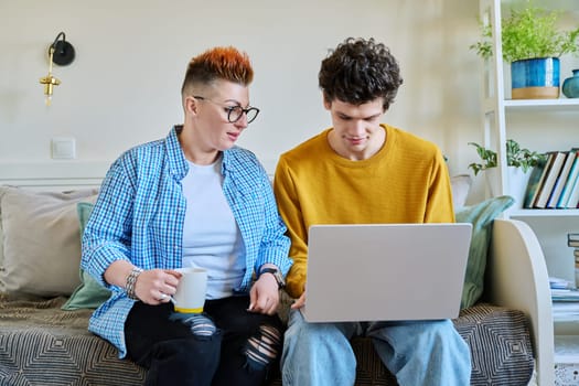 Middle-aged mother and son 19-20 years old sitting together on couch at home looking at laptop screen talking. Lifestyle family communication parenthood tenderness positive relationship two generation