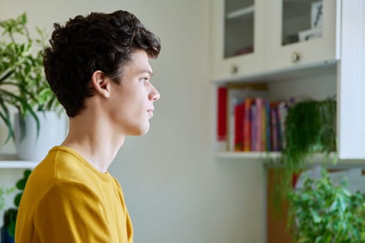 Profile portrait of young handsome guy with crossed arms, in home interior. Confident serious male 19-20 years old in casual yellow with curly hairstyle looking at window. Lifestyle, youth concept