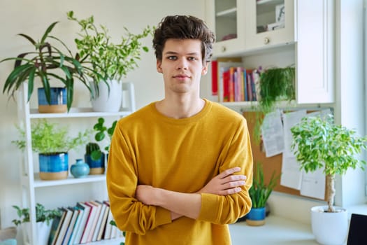 Portrait of young handsome guy with crossed arms, in home interior. Smiling confident male 19-20 years old in casual yellow with curly hairstyle looking at camera. Lifestyle, youth concept