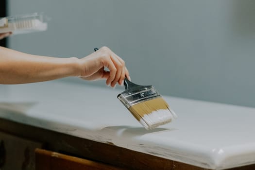 Young Caucasian unrecognizable girl paints an old wardrobe in her room with a brush and white paint, close-up side view.