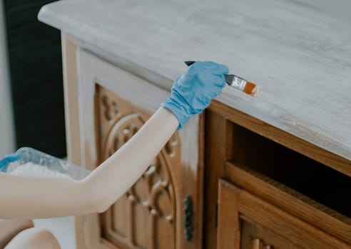 One young Caucasian unrecognizable girl in blue disposable gloves paints the patterned door of an old cabinet with white paint with a narrow brush, sitting in the room during the day, close-up top view.
