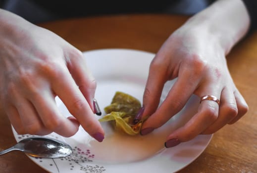 The hands of a young woman roll up a raw sauerkraut leaf with meat and rice filling in a plate while sitting at a round table in a flat kitchen. The concept of step by step instructions, home cooking, traditional recipes, national cuisine, cabbage rolls, dolma.