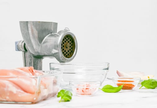 Manual retro meat grinder with glass bowl,boneless chicken thighs,pink salt,peeled onions,garlic in a saucer and basil leaves on a marble table against a white wall,closeup side view.Concept of home cooking and healthy eating.