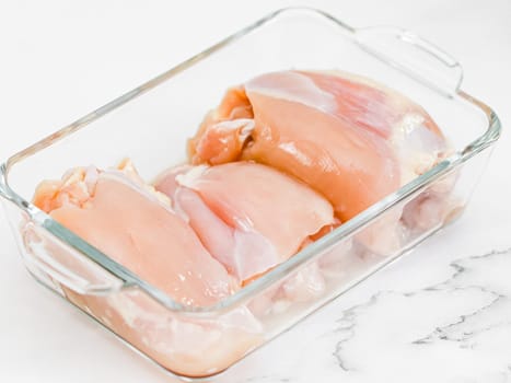 Raw boneless chicken thigh meat lies in a glass baking dish on a white marble table, close-up side view. Concept of raw chicken meat, raw food.