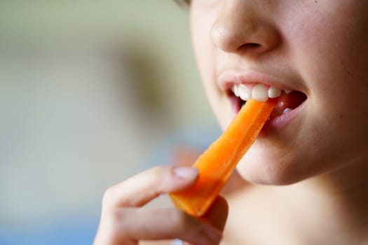 Closeup of anonymous crop young girl eating fresh organic carrot slice at home