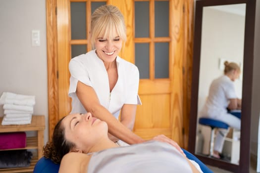 Positive female osteopath in uniform manipulating head of patient lying on couch during psychotherapy session in physical therapy clinic