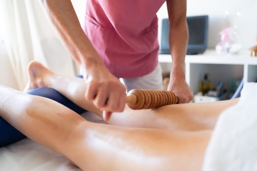 Crop anonymous female massage therapist massaging with wooden device on leg of patient lying on bed during rehabilitation session in modern clinic