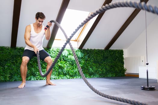 Full body of young fit male athlete in sportswear exercising with battle ropes while improving core strength power and endurance in modern gym