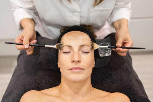 From above crop beauty specialist applying liquid cosmetic with brushes on face of female customer in modern salon during skin care treatment