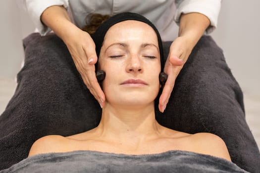 High angle of crop anonymous female cosmetician massaging cheeks of woman with natural crystals while smoothing facial skin