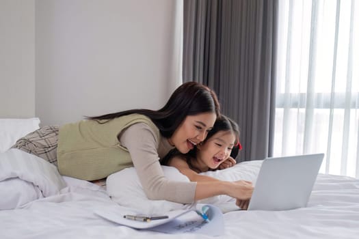 A young woman is working from home with her adorable daughter, watching work together and happily teasing each other in the bedroom..