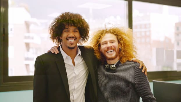 Two coworkers with curly hair embracing and smiling at camera in the office
