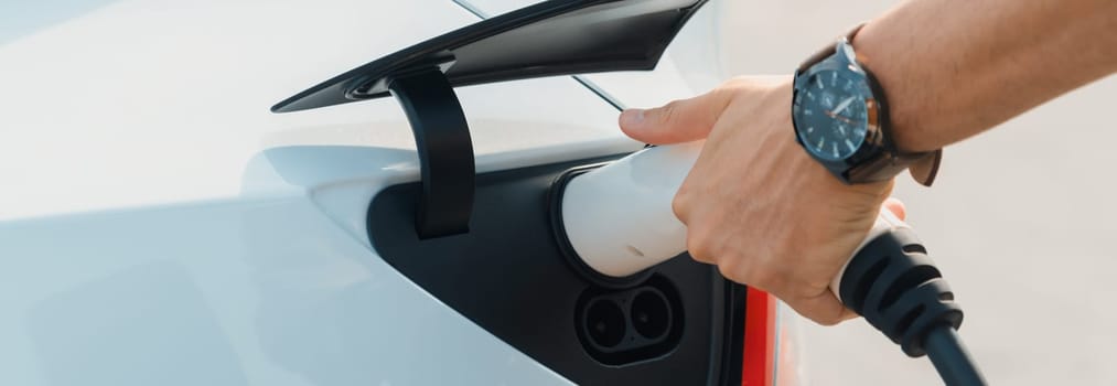 Closeup hand put EV charger to recharge electric car's battery in city commercial parking lot. Rechargeable EV car for sustainable environmental friendly urban travel. Panorama Expedient