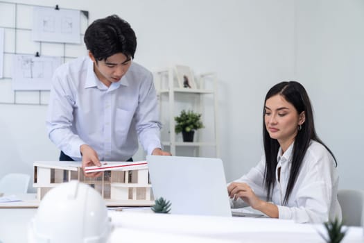 Professional male asian architect using ruler to measure house model length while young beautiful caucasian colleague using laptop to analyzed data on meeting table with house model. Immaculate.