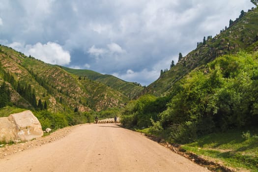 Dry dusty dirt road winding through valley with mountains, two shepherds lead a flock of sheep along the road at sunny day.