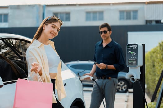 Young couple holding shopping bag and use smartphone to pay for electricity for recharging EV car battery from charging station at city mall parking lot. Happy couple go shopping by eco car. Expedient