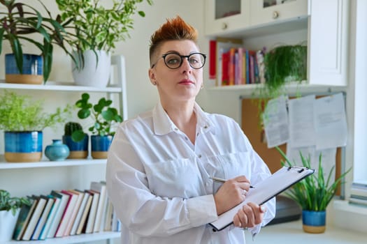 Middle aged confident successful business woman with clipboard at workplace. Remote business, teaching, work concept