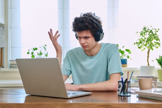 Young male student in headphones sitting at home at desk looking talking in web camera of computer laptop. Guy 19-20 years old studying remotely, video conference call, e-learning technology education