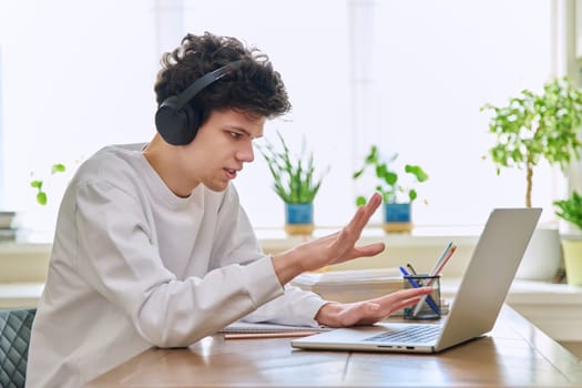 Young male student in headphones sitting at home at desk looking talking in web camera of computer laptop. Guy 19-20 years old studying remotely, video conference call, e-learning technology education