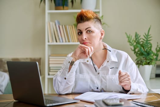 Portrait of mature businesswoman working at home on computer laptop. Serious tired middle-aged female holding glasses in hands looking at camera. Remote business work career management marketing