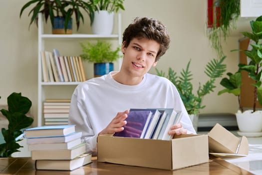 Satisfied young male customer buyer sitting at home unpacking cardboard box parcel with new books, online purchases. Delivery by mail, internet store bookstore