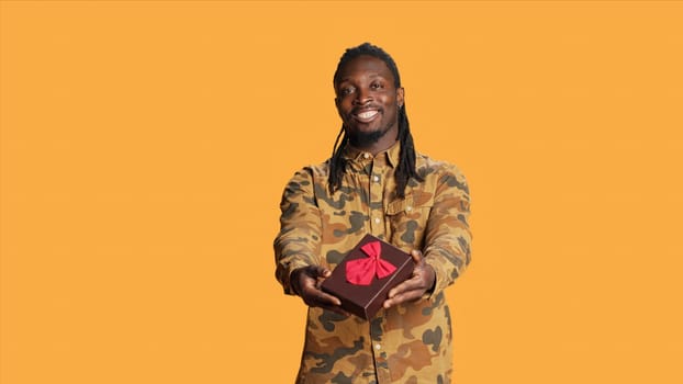 African american person offering giftbox to his girlfriend, giving present packed with ribbon and bow on camera. Young adult doing cute romantic gesture on their anniversary or valentines day.