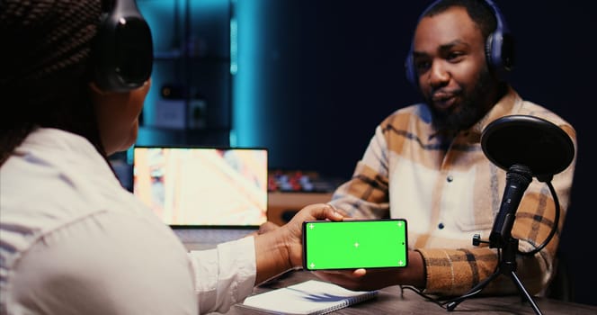 Man invited to podcast watching internet clips on green screen smartphone with host in studio, discussing content. BIPOC guest reacting to videos on mockup mobile phone during online comedy show