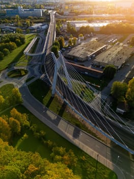 Aerial drone photo of modern suspension bridge in Krakow, Poland. Summer overhead view of buildings in sunlight, city park and high steel structure of bridge, road connection and intersection