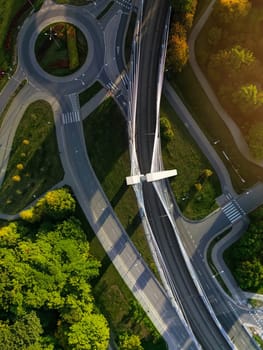 Aerial view directly from above of road ring and suspension bridge in Krakow, Poland. Scenic sunlight and shadows on treetops of city park, empty street without cars and modern bridge for trams