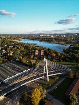 Aerial drone city view with suspension bridge in Krakow, Poland. Metal curved bridge structure, autumn park and houses of residential area, reflection of blue sky and clouds in water of Bagry bay