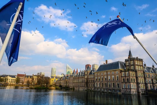 Waving NATO flag in the center of Hague sky with dutch river channels and old city buildings view. Holland sityscape,