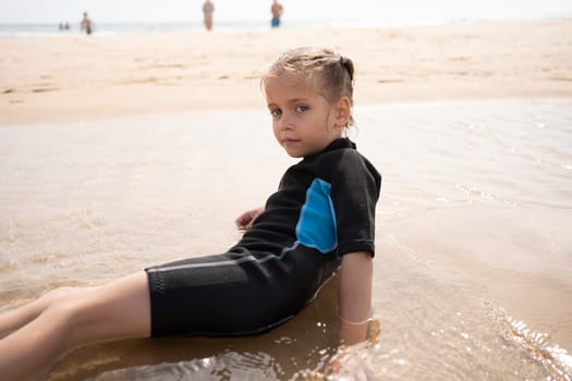 Female child surfer in wetsuit lies ocean beach. Serious child after surfing on sandy beach