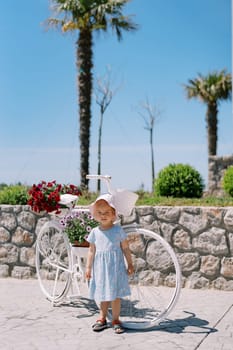 Little girl stands on the sidewalk near a white bicycle with potted flowers. High quality photo