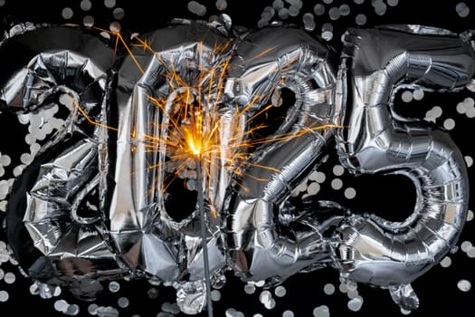 Happy new year 2025 metallic balloons with confetti and sparkler firework Bengal lights on dark black background. Greeting card silver foil balloons numbers Christmas holiday concept. Celebration party congratulation decoration