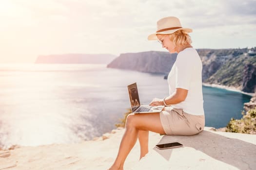 Successful business woman in yellow hat working on laptop by the sea. Pretty lady typing on computer at summer day outdoors. Freelance, travel and holidays concept.