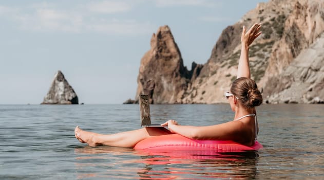 Woman freelancer works on laptop swimming in sea on pink inflatable ring. Pretty lady typing on computer while floating in the sea on inflatable donut at sunset. Freelance, remote work on vacation