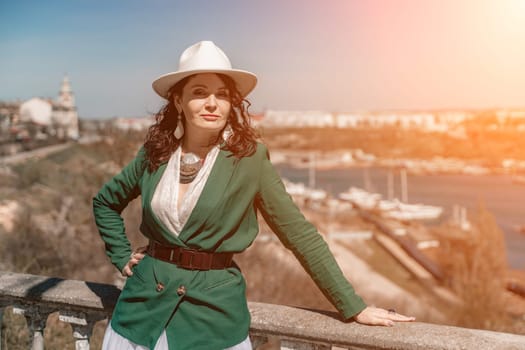 Woman walks around the city, lifestyle. Happy woman in a green jacket, white skirt and hat is sitting on a white fence with balusters overlooking the sea bay and the city