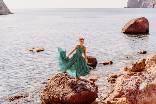 Woman green dress sea. Female dancer in a long mint dress posing on a beach with rocks on sunny day. Girl on the nature on blue sky background