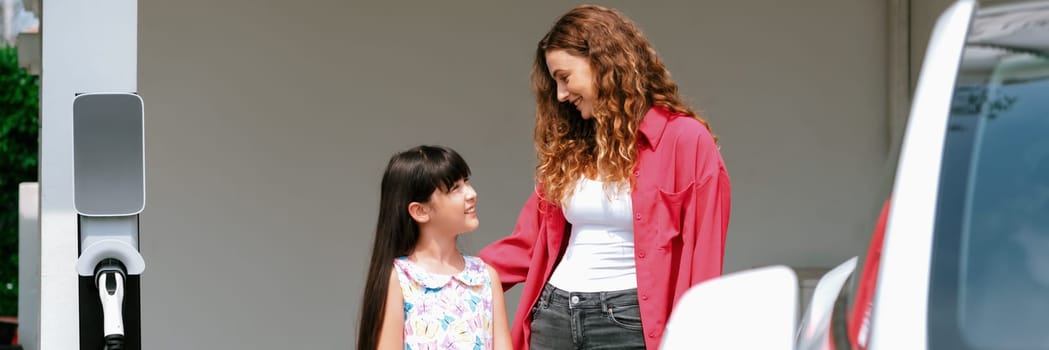 Happy little young girl learn about eco-friendly and energy sustainability as she help her mother recharge electric vehicle from home EV charging station. EV car and modern family. Panorama Synchronos