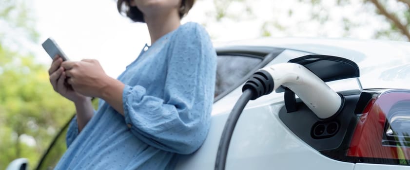 Focus EV charger by electric charging station recharging car on blurred background of beautiful young woman checking car's battery from smartphone. Eco-friendly travel with EV car. Panorama Perpetual