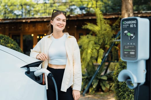 Sustainable urban commute with young woman pay electricity for EV electric car recharging at outdoor cafe in springtime garden, green city sustainability and environmental friendly EV car. Expedient