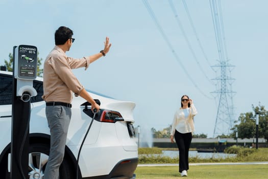 Young couple recharge EV car battery at charging station connected to power grid tower electrical industrial facility as electrical industry for eco friendly vehicle utilization. Expedient