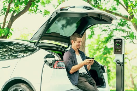 Man using smartphone online banking application to pay for electric car battery charging from EV charging station during vacation holiday road trip at national park or summer forest. Exalt