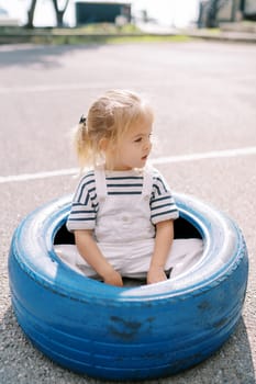 Little girl sits in a big blue tire on the playground and looks away. High quality photo