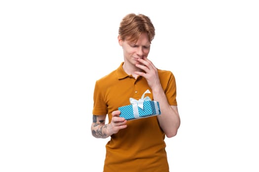young red-haired guy in an orange t-shirt holding a gift box on a white background with copy space.