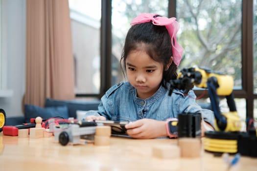 A young Asian girl in a STEM class attentively uses a smartphone app to remotely control a toy car, showcasing tech education.