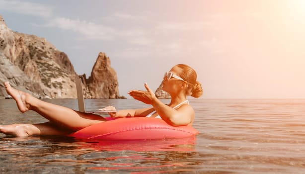 Woman freelancer works on laptop swimming in sea on pink inflatable ring. Pretty lady typing on computer while floating in the sea on inflatable donut at sunset. Freelance, remote work on vacation