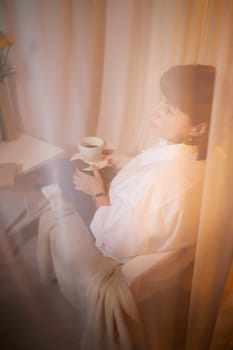 Nice girl with a mug of tea or coffee in cozy room. A middle-aged adult woman relaxing in the evening in the living room or bedroom in the evening